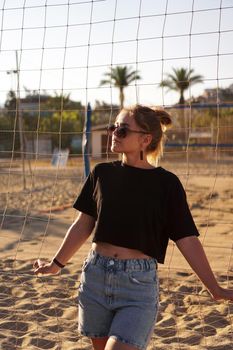 Portrait of attractive woman near volleyball net on the beach. Beautiful young woman in shorts, black tank top and sunglasses. Against the background of the beach and palms. Vertical photo