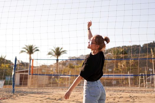 Portrait of attractive woman near volleyball net on the beach. Beautiful young woman in shorts, black tank top and sunglasses. Against the background of the beach and palms