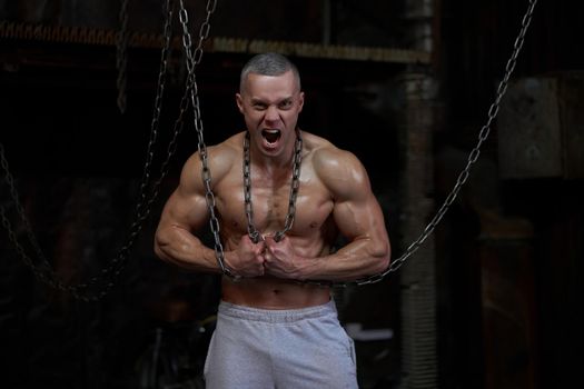Primal instinct. Angry muscular man screaming at camera and breaking chains on chest, dark background