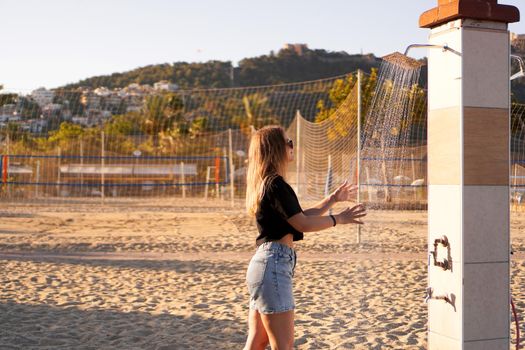 A girl in shorts and a black T-shirt on the beach near the shower. Shower on the beach. Nearby there are palm trees and a sports volleyball court.