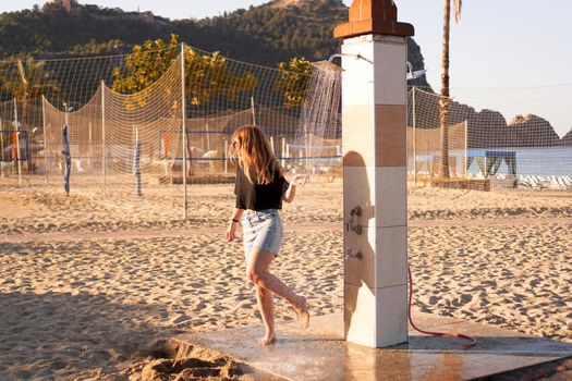 A girl in shorts and a black T-shirt on the beach near the shower. Shower on the beach. Nearby there are palm trees and a sports volleyball court.