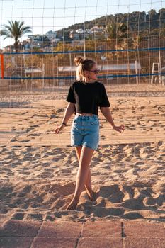 Portrait of attractive woman near volleyball net on the beach. Beautiful young woman in shorts, black tank top and sunglasses. Against the background of the beach and palms. Vertical photo