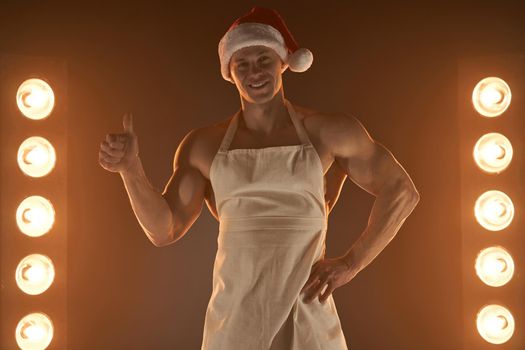 Hot Christmas dessert Sexy muscular Chef wearing apron and Santa hat thumb up gesture smiling, lamps illumination and smoke on background cuisine. male housewife. husband in kitchen. brutal butcher.
