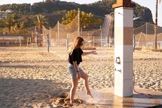 A girl in shorts and a black T-shirt on the beach near the shower. Shower on the beach. Nearby there are palm trees and a sports volleyball court.