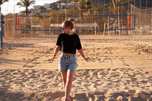 Portrait of attractive woman near volleyball net on the beach. Beautiful young woman in shorts, black tank top and sunglasses. Against the background of the beach and palms