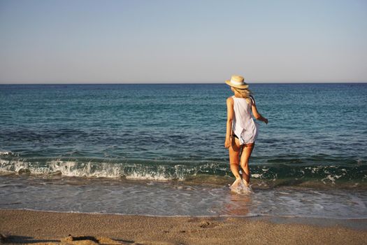 The girl walks on the beach. Beautiful blonde woman in white blouse and straw hat enjoying the sea