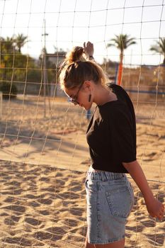 Portrait of attractive woman near volleyball net on the beach. Beautiful young woman in shorts, black tank top and sunglasses. Against the background of the beach and palms. Vertical photo