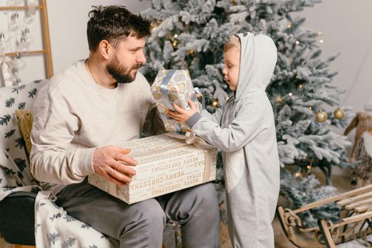 Holiday Christmas Handsome father playing with small cute son near decorated New year tree at home Family tradition boy gives a gift to his father