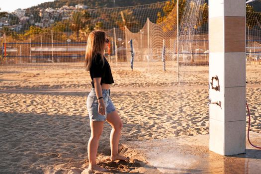 A girl in shorts and a black T-shirt on the beach near the shower. Shower on the beach. Nearby there are palm trees and a sports volleyball court.