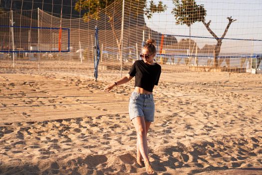 Portrait of attractive woman near volleyball net on the beach. Beautiful young woman in shorts, black tank top and sunglasses. Against the background of the beach and palms