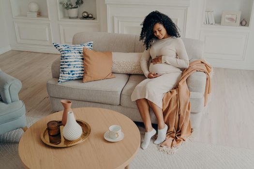 Beautiful photo of pregnant afro american lady sitting on cozy sofa decorated with colorful pillows gently placing hands on her stomach while relaxing of living room at home, enjoying pregnancy time