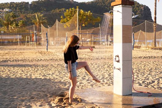 A girl in shorts and a black T-shirt on the beach near the shower. Shower on the beach. Nearby there are palm trees and a sports volleyball court.