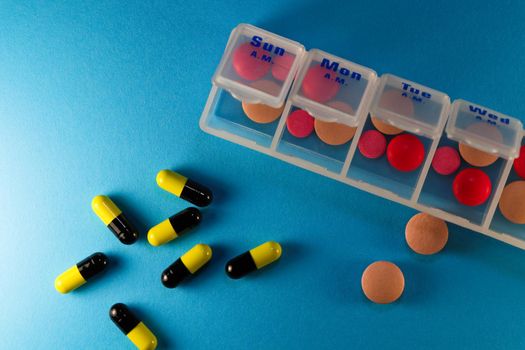 Various pills and capsules with a daily prescription drug sorting container on blue background abstract flat-lay