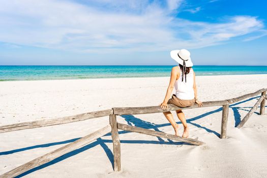 Young woman on vacation alone admires the crystal clear tropical sea sitting on a wooden fence on a white sand beach under blue sky. Pensive girl with large white hat enjoying summer travel