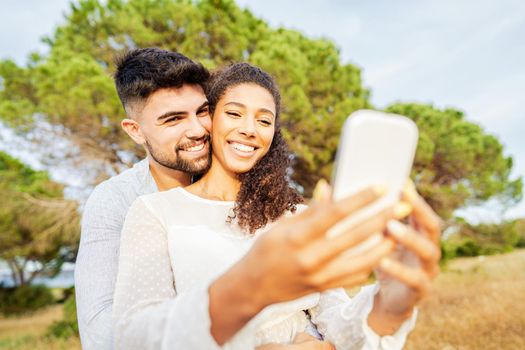 Young beautiful multiracial couple in love taking selfie in nature enjoying vacation moments sharing photos on social networks. New normal technology addiction due to wi-fi internet mobile connections