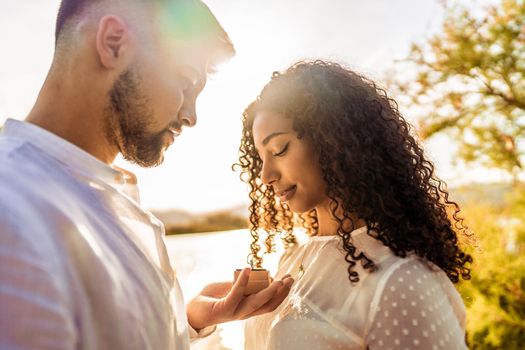 Backlit suggestive romance scene of young beautiful multiracial couple in love. Handsome guy making the marriage proposal to her black Hispanic girlfriend showing engagement ring close to her