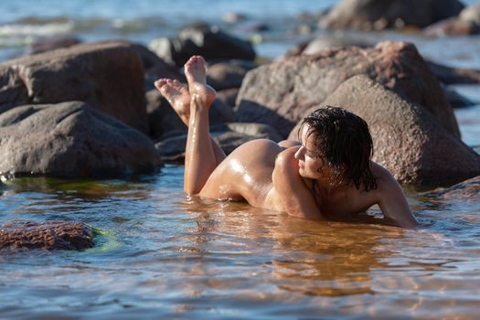 Nudity and a healthy lifestyle. Young nude woman with small breasts enjoys holiday at the seaside. Young nude woman lying in water on beach