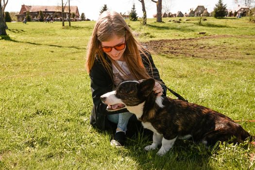 Young woman in red sunglasses plays with her corgi on a summer green lawn. Sunny day. Happy pet and owner