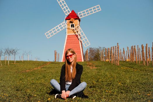 Beautiful young woman in red sunglasses sits on the grass against the backdrop of the red mill. Sunny summer day. Rest in the countryside