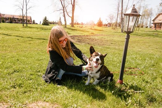 Young woman in red sunglasses plays with her corgi on a summer green lawn. Sunny day. Happy pet and owner