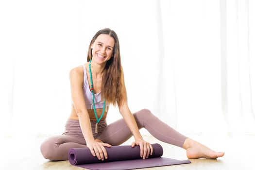 Young sporty female yoga instructor in bright white yoga studio, smiling cheerfully while rolling the yoga mat after yoga session. Smiling, looking at camera.