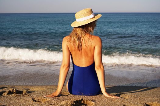 Young Woman Sunbathing At Tropical Beach. Woman in a blue swimsuit and hat sits on the seashore on sand