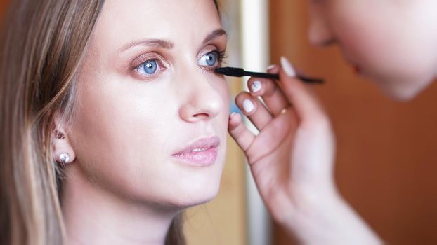 Process of making makeup. Make-up artist working with brush on model face. Portrait of young woman in home interior.