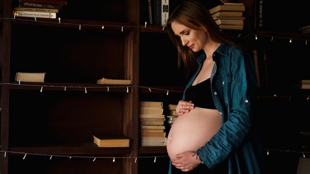 Beautiful young stylish pregnant woman, in black lingerie and blue shirt, in the interior of a loft on the background of wooden bookcase with books