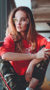 Fashion model wearing red hoodie with the inscription los angeles and big plastic glasses posing in the city at parking. Fashion urban outfit