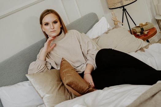 Woman with bright makeup and strict facial features dressed in beige sweater sitting on bed