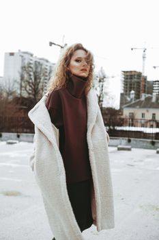 A girl with red curly hair in a white coat poses on outdoor parking in cold autumn. City Style - Urban. vertical photo