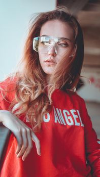 Fashion model wearing red hoodie with the inscription los angeles and big plastic glasses posing in the city at parking. Fashion urban outfit