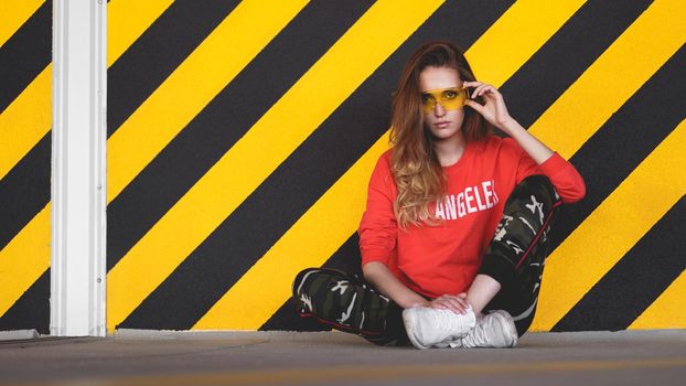 Fashion model wearing red hoodie with the inscription los angeles and big plastic glasses posing in the city at parking. Fashion urban outfit