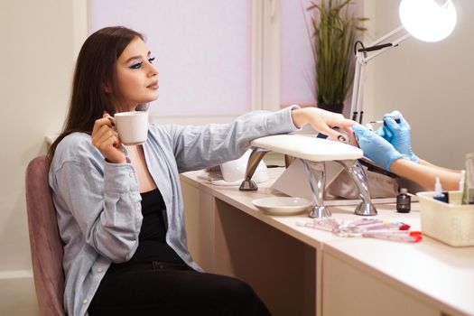 Beautiful woman getting a manicure in a beauty salon. She smiles and holds a white cup of coffee in her hand.