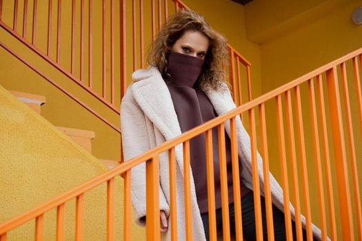 A girl with red curly hair in a white coat poses on the yellow orange parking stairs. City Style - Urban