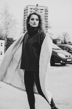 A girl with red curly hair in a white coat poses on outdoor parking in cold autumn. City Style - Urban