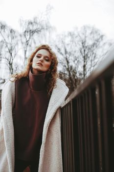 A girl with red curly hair in a white coat poses on outdoor parking in cold autumn. City Style - Urban