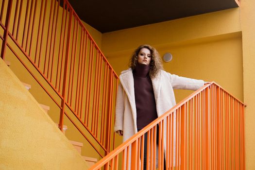 A girl with red curly hair in a white coat poses on the yellow orange parking stairs. City Style - Urban