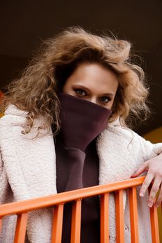 A girl with red curly hair in a white coat poses on the yellow orange parking stairs. City Style - Urban