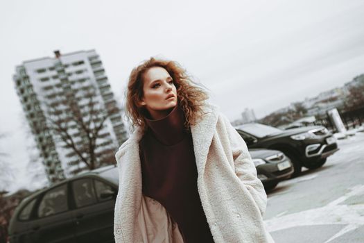 A girl with red curly hair in a white coat poses on outdoor parking in cold autumn. City Style - Urban