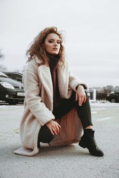 A girl with red curly hair in a white coat poses on outdoor parking in cold autumn. City Style - Urban