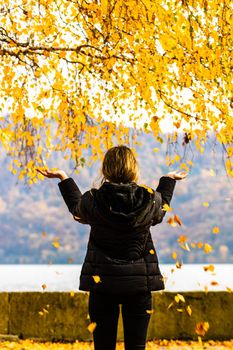 Back view of alone woman enjoying autumn, throwing fallen leaves on autumn alley. Autumn landscape, orange foliage in a park in Orsova, Romania, 2020