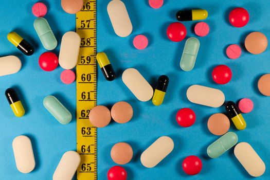 Various pills and capsules with measuring tape on blue background flat-lay weight-loss concept