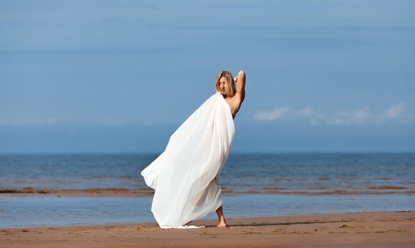 Young naked blonde posing on the sea coast on a summer sunny day with a white fabric flying in the wind. Sexy woman enjoying summer time