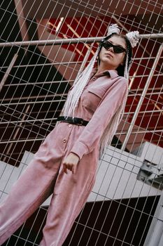Portrait of beautiful cool girl over urban wall. Sunny day. Girl in sunglasses and pink jumpsuit