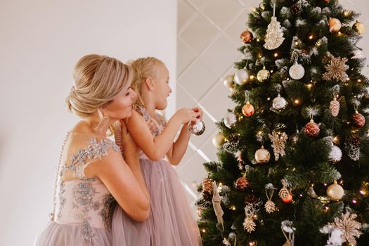 Young beautiful mother holds a little daughter who decorates a Christmas tree in the cozy room