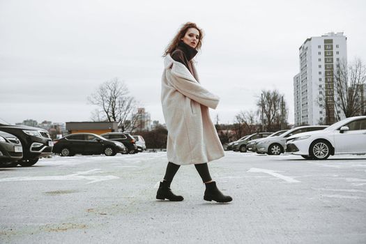 A girl with red curly hair in a white coat poses on outdoor parking in cold autumn. City Style - Urban