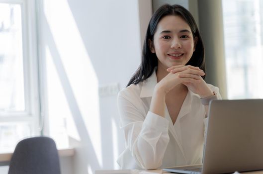 Beautiful Asian business lady is looking at camera and smiling while working laptop in bright modern office with copy space.