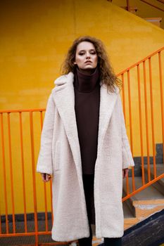 A girl with red curly hair in a white coat poses on the yellow orange parking stairs. City Style - Urban