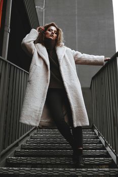 A girl with red curly hair in a white coat poses on the parking stairs. City Style - Urban